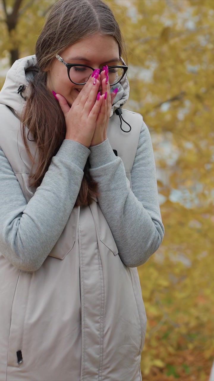Buitenbeeld toont herfstbladeren en een gedeeltelijk uitzicht. Fijne details zijn onder andere lang haar, een bril en een vest over een hoodie. De parkachtige omgeving met gele bladeren blijft onveranderd.