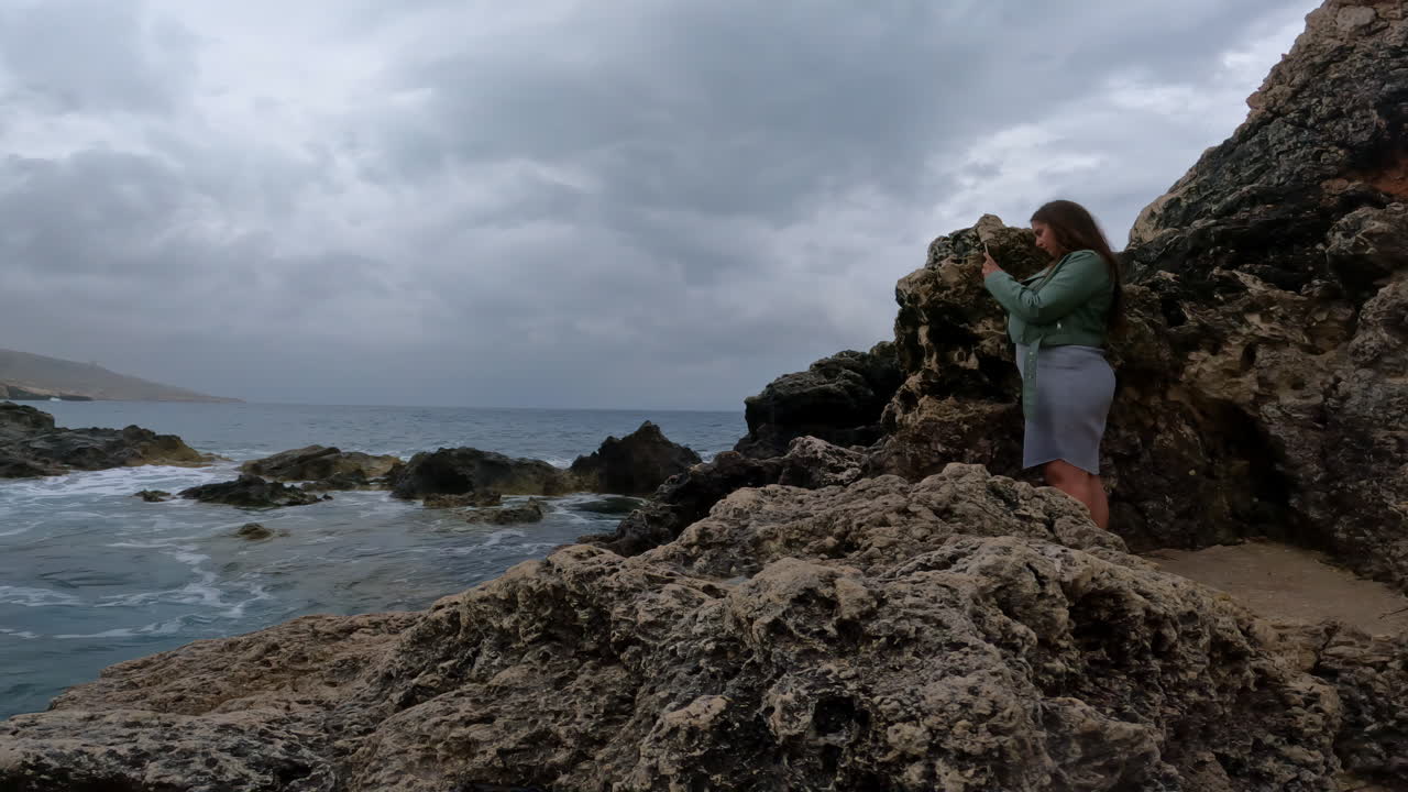 medio portátil filmado en la costa de malta con una vista de una influyente viajera fotografiando las rocas y las olas del mar para su publicación en las redes sociales en su emocionante viaje de vacaciones