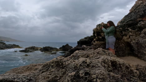 medium handheld shot off the coast of malta with a view of a traveling influencer photographing the rocks and waves of the sea for her social media post on her exciting vacation trip