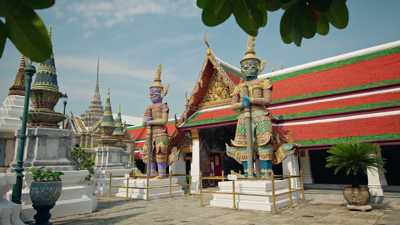 Estátuas gigantes e coloridas de guardiões no templo Wat Phra Kaew, em Bangkok, Tailândia, emolduradas por folhagem tropical.