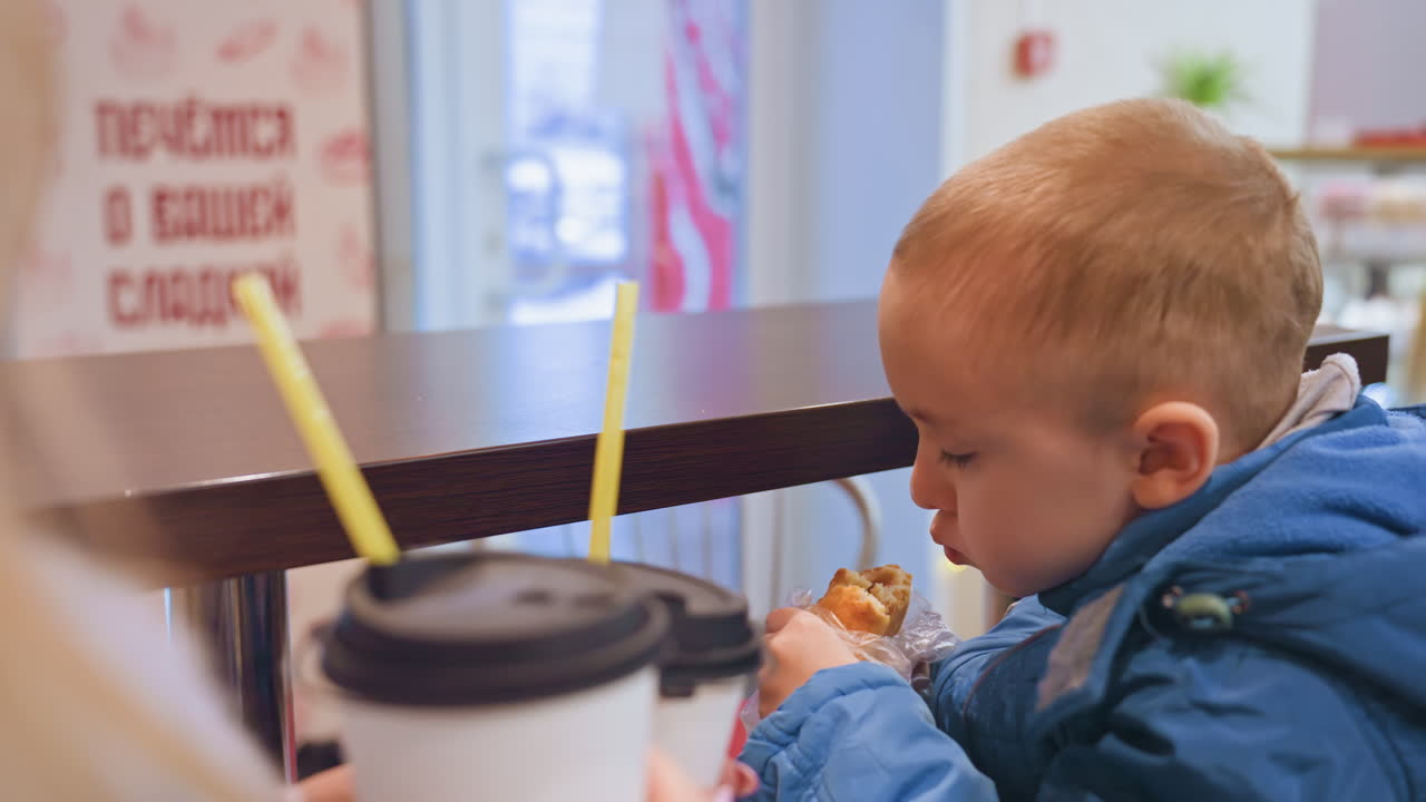 Mamma e figlio al bancone che condividono un dolce e un caffè caldo in un caffè interno, la madre tiene una tazza da asporto mentre il bambino con il cappotto blu esamina la cannuccia e morde il dolce, interazione delicata, illuminazione accogliente