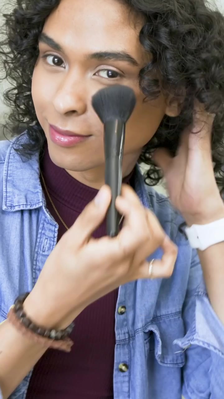 Vertical video: Leaning nonbinary maroon top checking mirror, blending with brush at sink, grooming