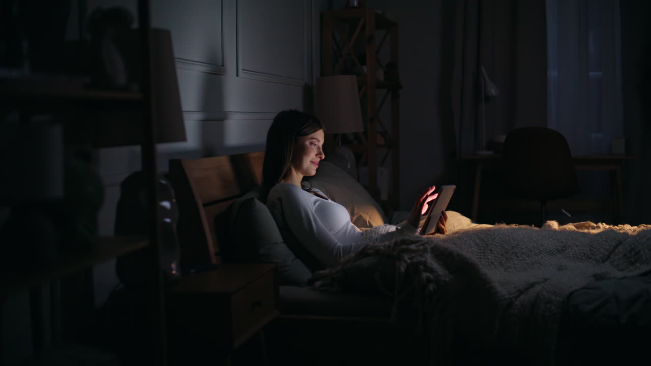 Donna a casa che tocca lo schermo luminoso del tablet sdraiata comodamente nel letto di casa di notte