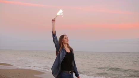 Mujer-Joven-Y-Atractiva-Parada-Junto-Al-Mar-Durante-La-Puesta-De-Sol-Y-Sosteniendo-Una-Vela-Brillante-Encendida.-Tiro-En-Cámara-Lenta