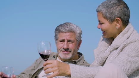 Cheerful-senior-wife-and-husband-sitting-on-shore-and-hugging