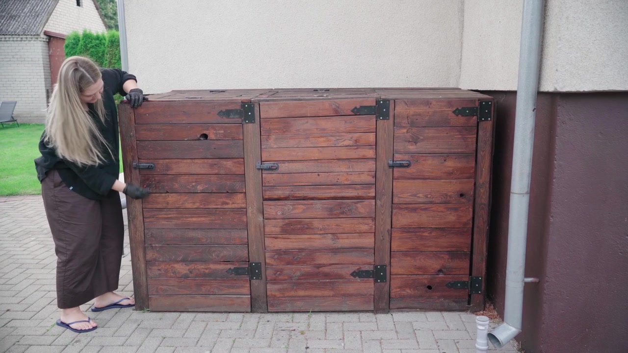 Woman in work gloves pushes against a poorly built wooden outdoor bin enclosure, checking its loose panels and hardware.