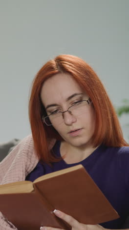 redhead woman in glasses enjoys reading captivating love story in evening. focused lady sits on sofa near cup of tea on special folding table at home