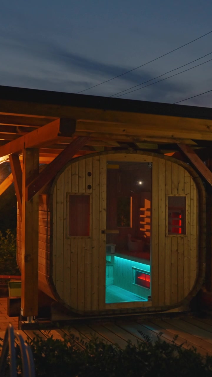 Una moderna sauna a botte risplende in modo invitante con vivaci luci interne in questo video verticale, perfetto per dispositivi mobili e social media, ripreso al tramonto all'aperto