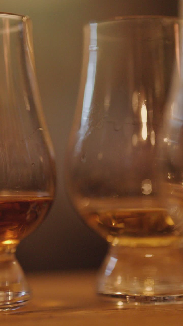 Una fluida panoramica della telecamera rivela bicchieri da whisky con liquido ambrato su un bancone di legno, sotto una calda illuminazione ambientale in un accogliente ambiente da sala degustazione.