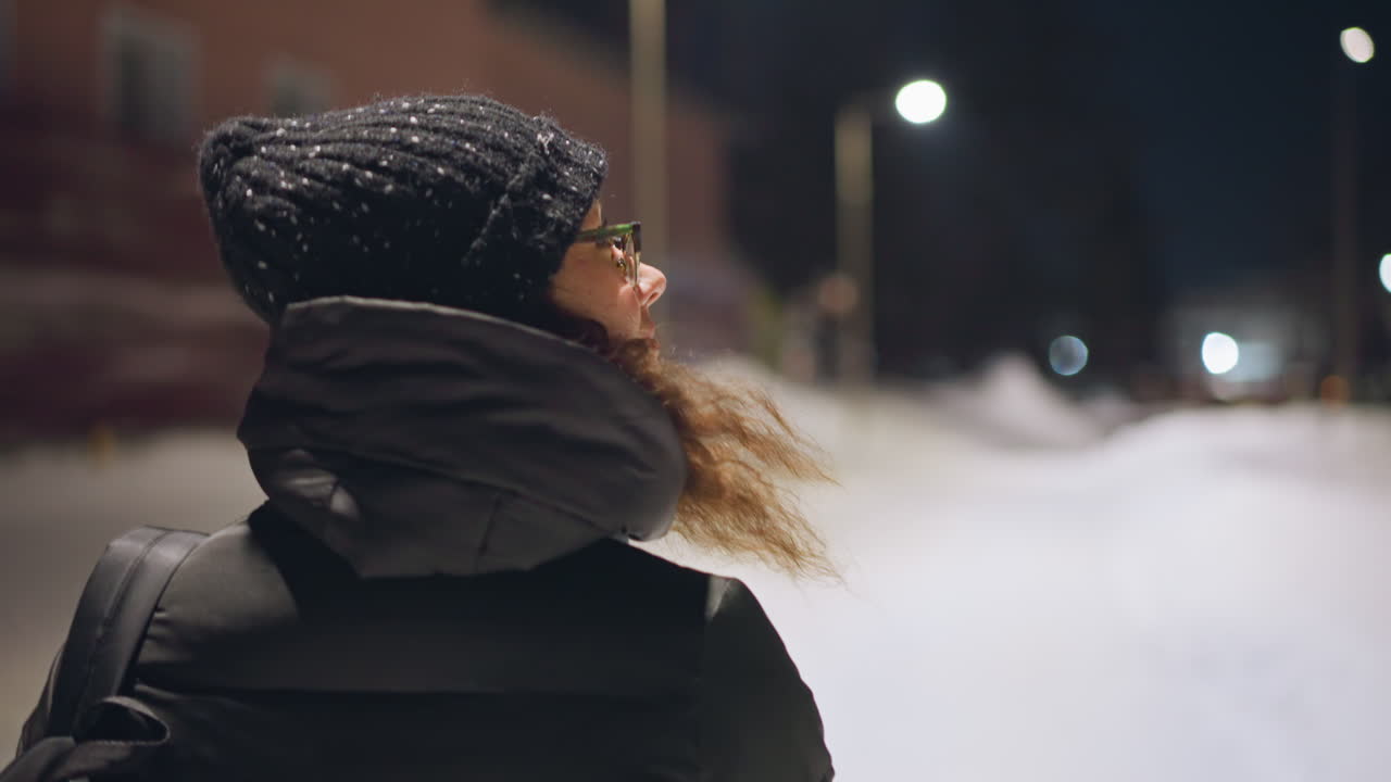 Woman in warm winter coat and knitted hat walking outside at night on snowy street, wearing glasses and colorful gloves, illuminated by streetlights, enjoying cold evening atmosphere in peaceful quiet urban