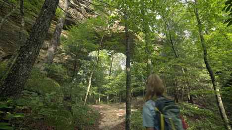 female hiker walks under natural stone arch formation among lush trees, 4k