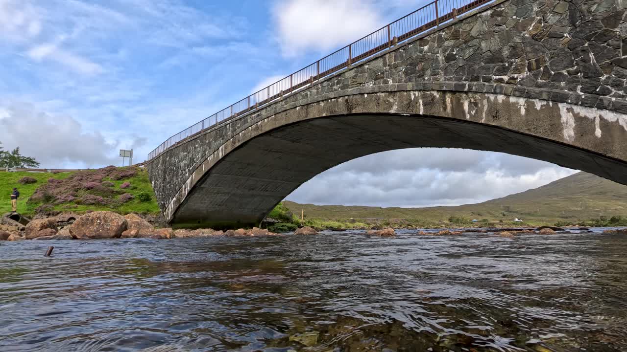 Camera transitions from underwater stones to a wide, low-angle view of a stone bridge over a flowing stream, with natural daylight and gentle movement