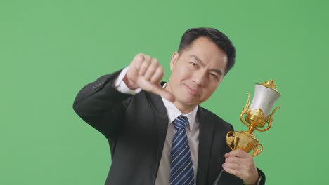 close up of asian business man in a suit and tie with a gold medal and trophy showing thumbs down gesture to ridicule the loser on green screen background in the studio