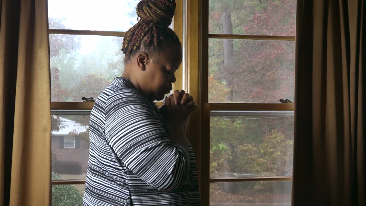 Black woman in her 40's standing by window on a rainy day praying to God