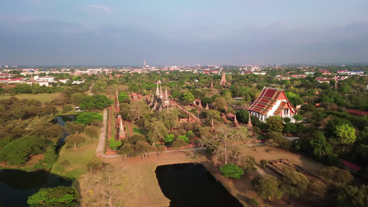 Aerial approach over ponds to temple ruins in Ayutthaya with flat surrounding plains and distant city line