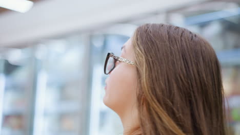 primer plano de una mujer caminando en un centro comercial bien iluminado, mirando a su alrededor contemplativamente a las mercancías expuestas detrás del vidrio, la luz suave se refleja en la pantalla