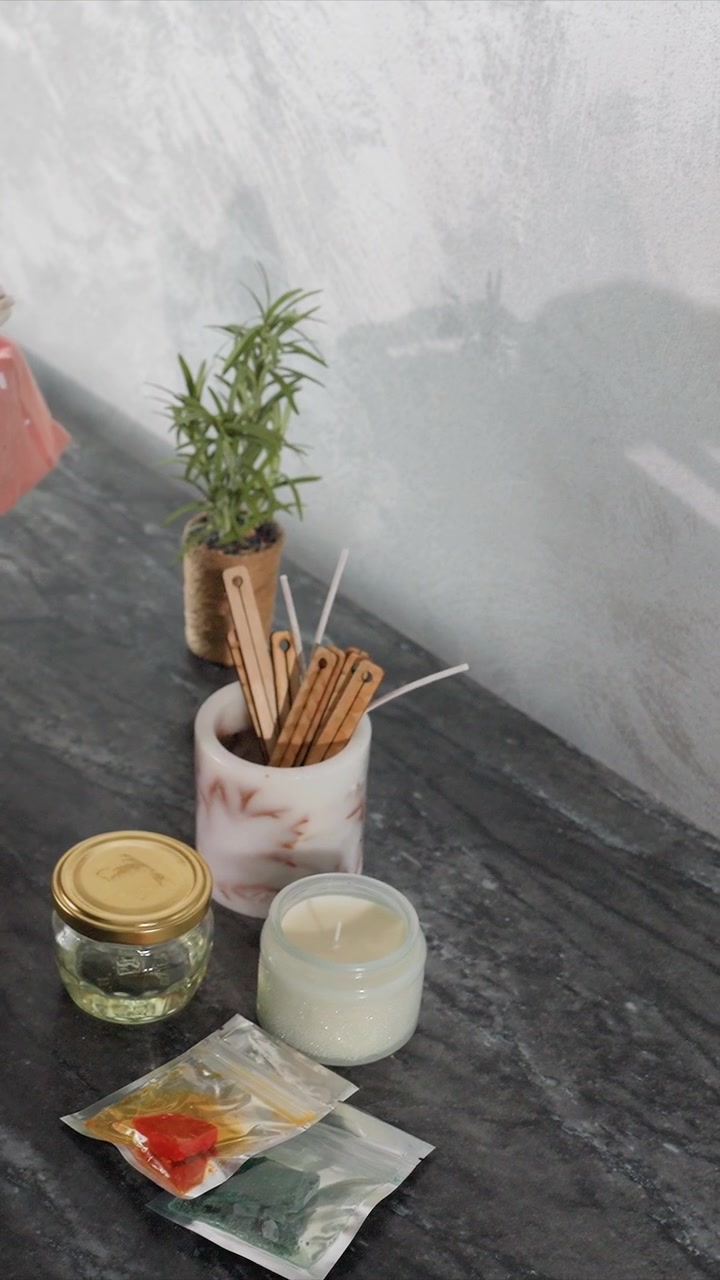 Gros plan sur une personne arrangeant des matériaux de fabrication de bougies sur une surface en bois foncé, notamment un pot en verre avec un couvercle doré et un récipient de cire blanche, placés à côté d'une petite plante en pot.