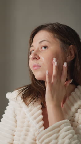 dama usa cosméticos para la cara de pie en el espejo. dama morena con cabello corto y húmedo en túnica de peluche aplica gel hidratante a la cara en un lugar claro