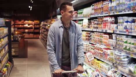 sonriente, hombre elegante con un carrito de compras en un gran centro comercial caminando por los estantes con productos lácteos y de repente comienza