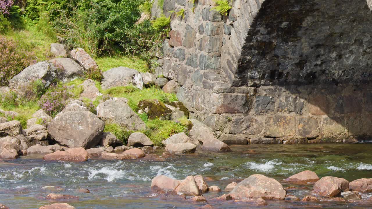 Camera pans across historic stone bridge and river, bright daylight, lush green landscape, tranquil mood