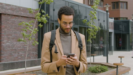 smiling american man in formal clothes using mobile phone and looking around while standing with his bike in the street