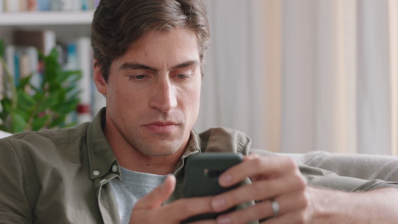 스마트 폰을 사용하는 매력적인 젊은 남자가 문자 메시지를 공유하고 휴대 전화로 온라인 소셜 미디어를 탐색하고 집에서 휴식을 취하고 있습니다 4k 영상