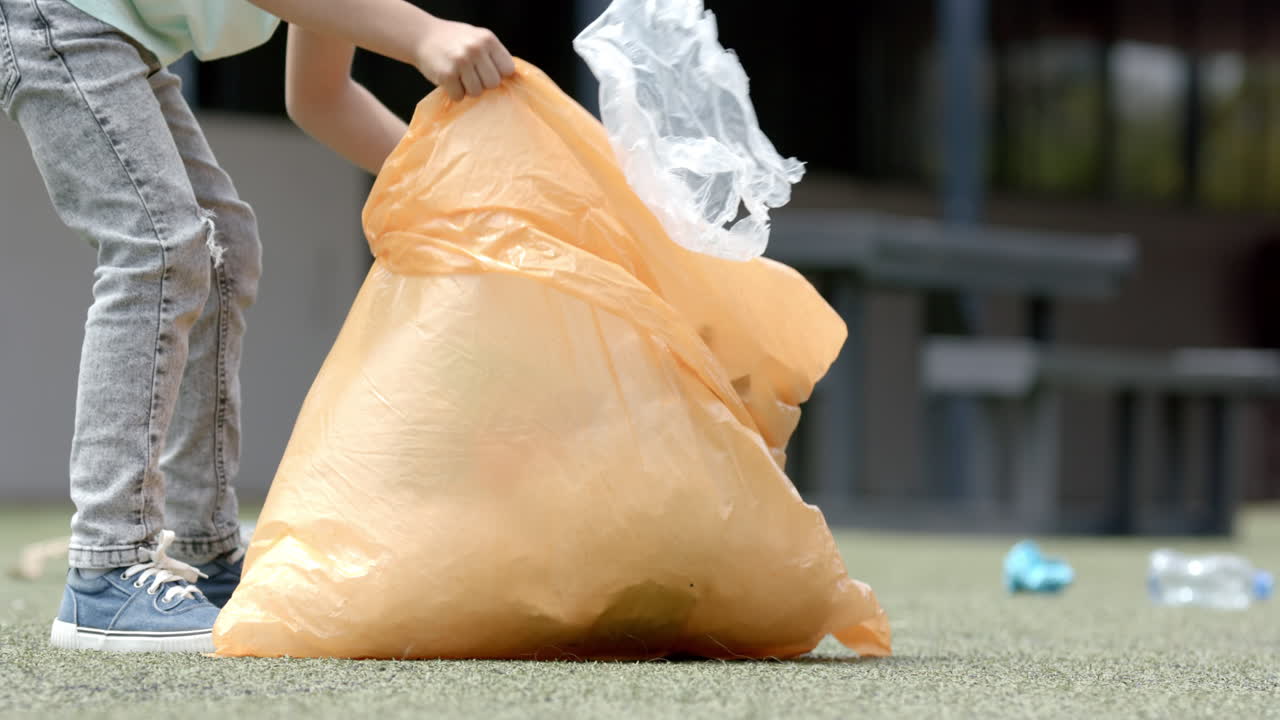 In school, child picking up trash with large orange bag, cleaning playground