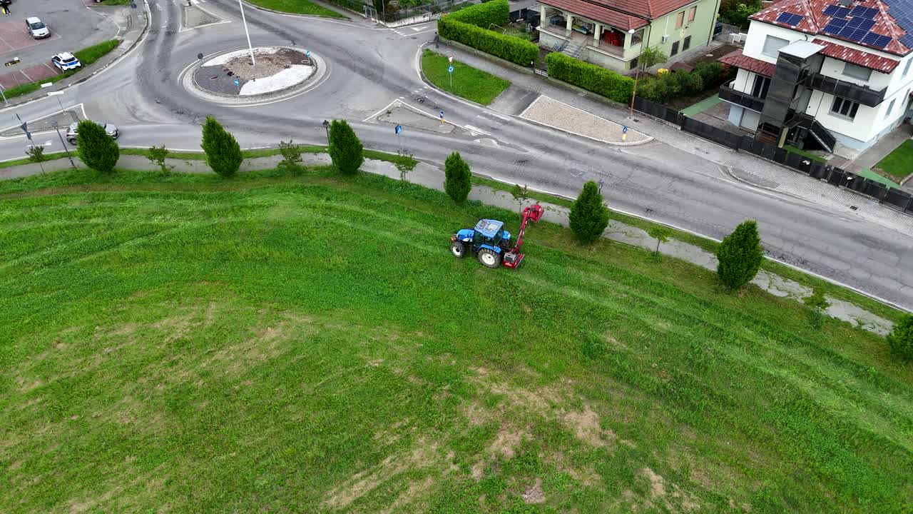 Agricultural worker riding tractor, cutting grass near roundabout, blending farming activity with residential suburban setting in Piacenza, northern Italy