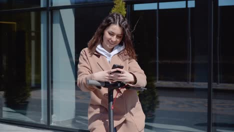 a young student girl in a sweater, jeans and a coat stands with an electric scooter near a stylish modern building and types in a smartphone. front view