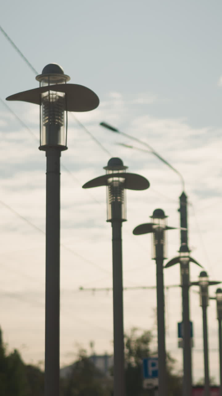 Farolas altas que bordean un bulevar tranquilo, retroiluminación del atardecer que proyecta largas siluetas y un brillo cálido, cables aéreos y postes que retroceden creando una perspectiva rítmica, tonos cinematográficos suaves que evocan recuerdos