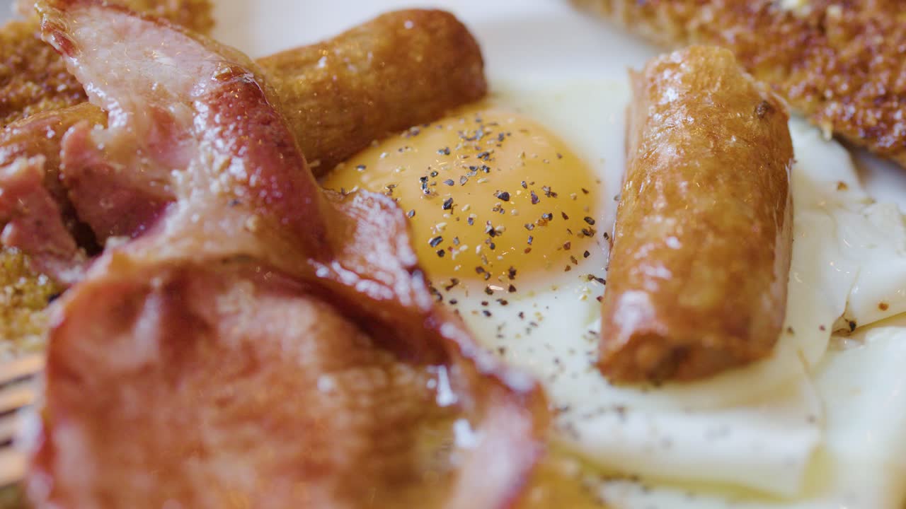 Close-up of fork placing bacon on eggs, sausage, toast, and beans under natural lighting