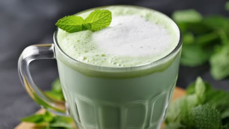 matcha latte decorated with mint leaves is steaming inside a transparent glass cup with handle, placed on a dark background, near other fresh mint leaves