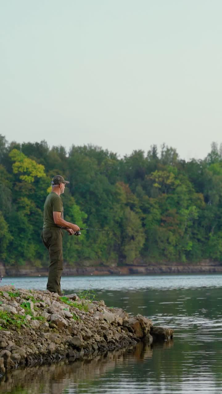 Vertical video of active senior fishing along the river coast with verdant forest in the background. Enjoying retirement in nature.