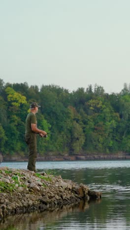 vertical video of active senior fishing along the river coast with verdant forest in the background. enjoying retirement in nature.