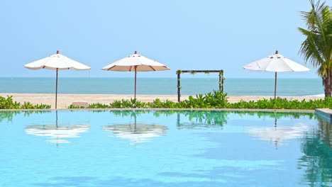 empty pool and sandy beach on tropical island resort, umbrellas water reflection and green vegetation, full frame