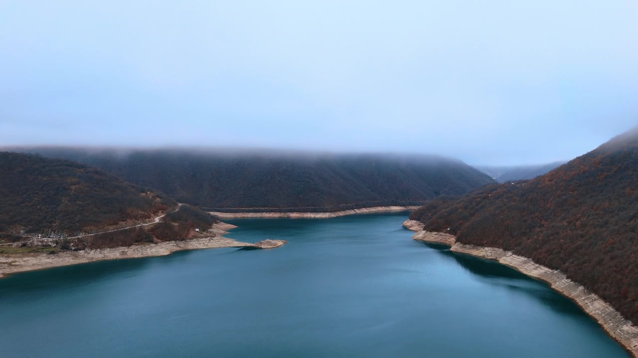 Symmetric view of wide mountain reservoir between fog covered hills. Fog covered hills flank a wide teal reservoir and low water levels expose broad white sediment banks along both shores.