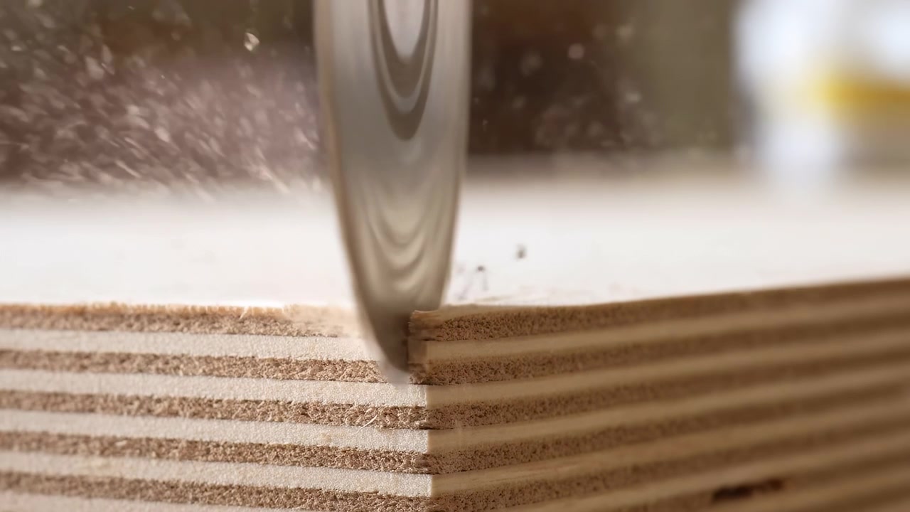 Vista macro ravvicinata di una lama di sega circolare che taglia attivamente pannelli di compensato impilati, con separazione di impiallacciatura a strati, polvere di legno, trucioli, contatto metallico affilato e texture superficiale visibile che crea un forte