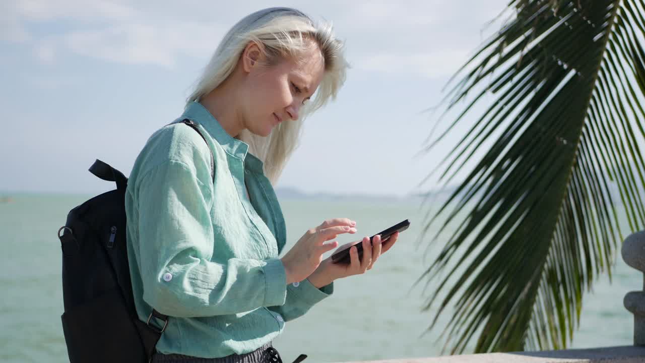 hermosa mujer delgada con cabello rubio largo y camisa verde de pie cerca de la palmera y usando el teléfono inteligente sobre el fondo del mar. niña pantalla táctil