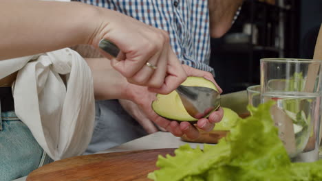 primo piano di una donna mani che scavano la carne fuori dall'avocado, mentre l'uomo affetta l'avocado in un tagliere di legno in una cucina moderna