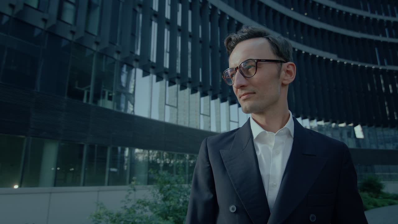 elegante hombre de negocios guapo en traje negro y gafas de pie al aire libre luego mirando a la cámara, retrato cinematográfico