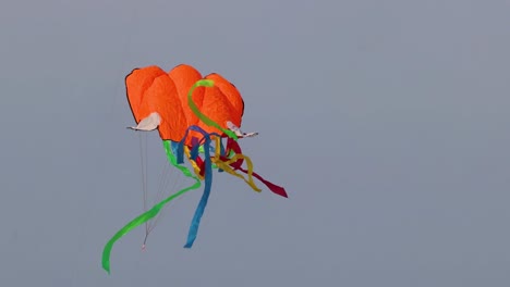 an orange kite with vibrant tails dances dynamically against a clear sky.