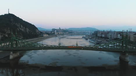 Aerial-view-of-yellow-tram-passing-through-Liberty-bridge-on-river-Danube-in-Budapest-at-sunrise,-morning