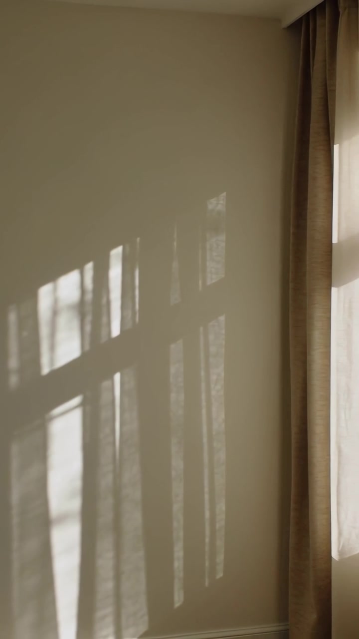 Vertical video: Sun via window casting muntin and tree shadows on cream wall at home by dual drapes