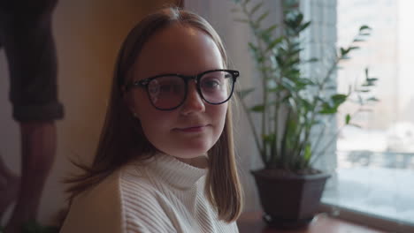 young woman in glasses seated at wooden table with warm mug in hand glancing toward camera, with indoor wall art, potted plant beside her, and snowy cityscape visible through large window behind