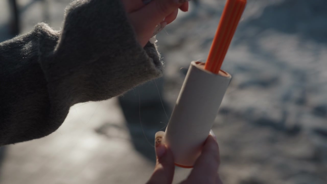 Primo piano di mani che staccano un rullo levapelucchi all'aperto, maniche di maglione grigio, unghie curate sollevano delicatamente la striscia adesiva, movimento tattile soddisfacente, sfondo sfocato di un marciapiede invernale, routine intima e accogliente.