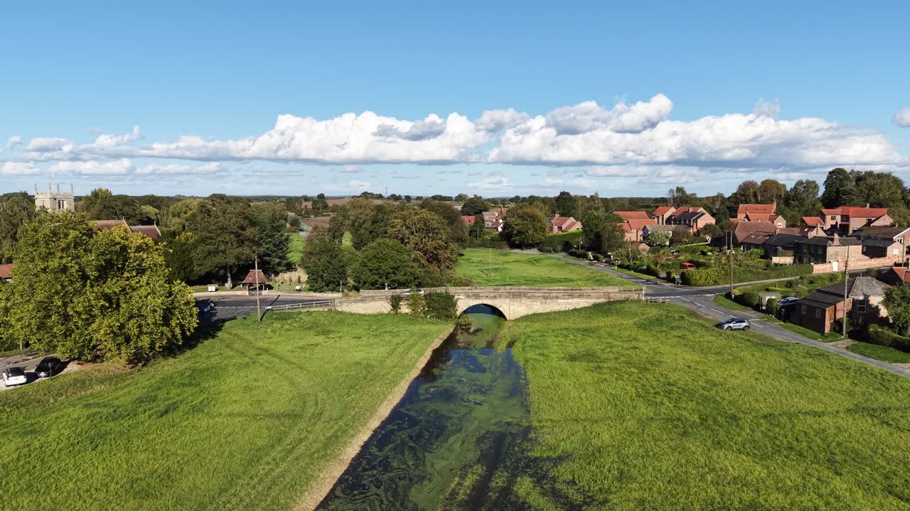 Dronekuva Thornton le Dalesta Pohjois-Yorkshiressa, Englannissa, näyttäen kylätaloja, kivisillan, vihreitä peltoja, joen ja maaseutua pilvisen taivaan alla.