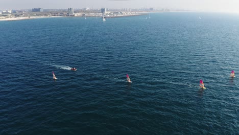 multiple windsurfers surf at high speed over the blue waters of the mediterranean sea with the coast of herzeliya in the background. high angle drone dolley shot