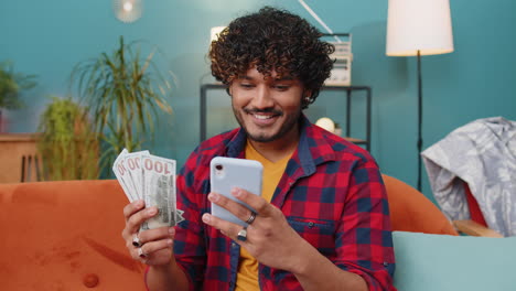 Smiling-happy-indian-man-counting-money-cash-use-smartphone,-income-saves-lottery-win-budget-at-home