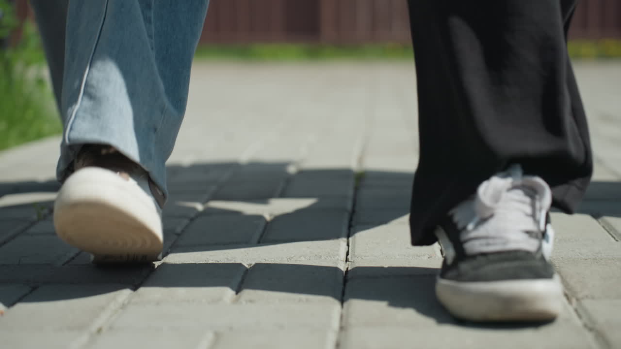 Close up of legs walking on paved courtyard, showing casual sneakers and denim in motion, casting long shadows on gray bricks under bright sunlight with hints of green grass at courtyard edge