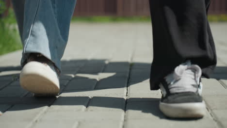 close up of legs walking on paved courtyard, showing casual sneakers and denim in motion, casting long shadows on gray bricks under bright sunlight with hints of green grass at courtyard edge
